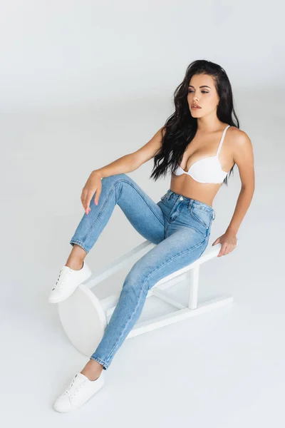 Брюнетка в джинсах и лифчике сидит на стуле на сером — стоковое фото