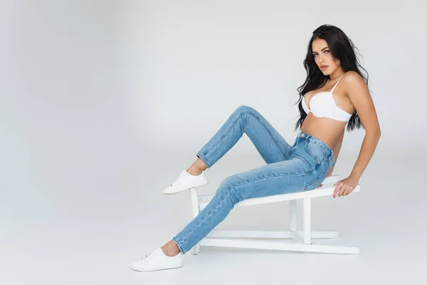 Sexy young woman in jeans and bra sitting on stool on grey — Stock Photo