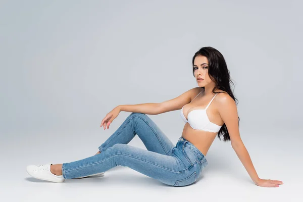 Jovem e morena mulher em sutiã e jeans olhando para a câmera enquanto sentado em cinza — Fotografia de Stock