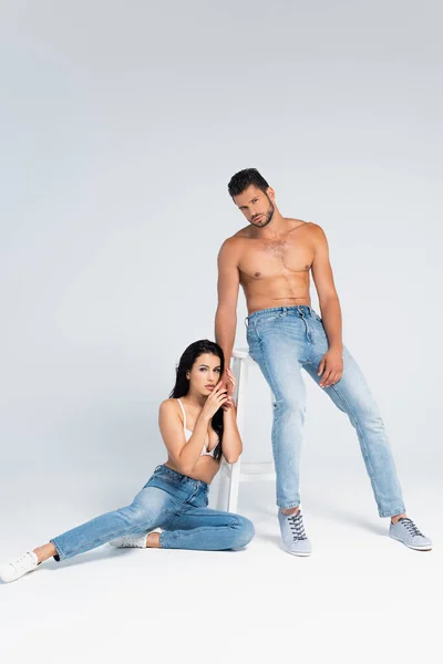 Femme sexy en soutien-gorge assis près de l'homme musclé et chaise sur gris — Photo de stock