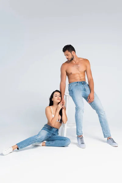 Sexy woman in bra sitting and holding hand of muscular man on grey — Stock Photo