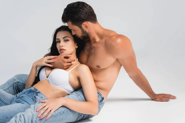 Muscular man in denim jeans hugging woman in bra lying on grey — Stock Photo