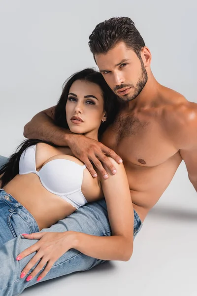 Muscular man in jeans hugging woman in white bra lying on grey — Stock Photo