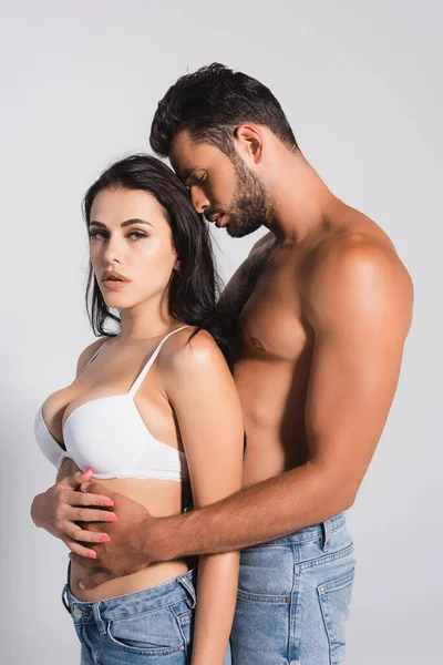 Uomo muscoloso in piedi e abbracciando donna sexy in reggiseno isolato su grigio — Foto stock