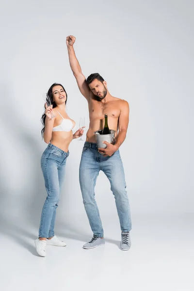 Muscular man holding ice bucket with bottle of champagne near sexy woman in bra standing with glasses on grey — Stock Photo