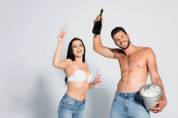 Мужчина без рубашки, держащий ведро со льдом и бутылку шампанского рядом с сексуальной женщиной в лифчике, стоящей в очках на сером — стоковое фото