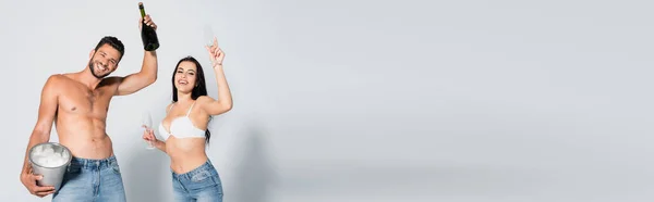 Récolte panoramique de muscle homme tenant seau à glace et bouteille de champagne près femme sexy en soutien-gorge debout avec des lunettes sur gris — Photo de stock