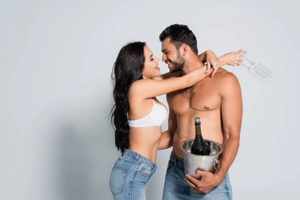 Mujer seductora en sujetador sosteniendo vasos y abrazando al hombre sin camisa con cubo de hielo y botella de champán en gris - foto de stock