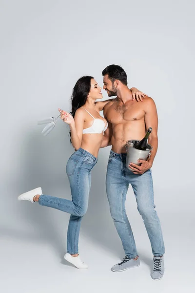 Muscular man holding ice bucket with bottle of champagne near seductive woman in bra with glasses standing on grey — Stock Photo