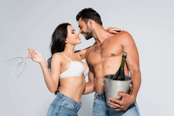 Hombre musculoso sosteniendo cubo de hielo con botella de champán cerca de mujer seductora en sujetador con gafas aisladas en gris - foto de stock