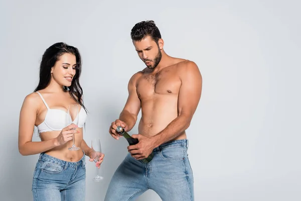 Мускулистый мужчина открывает бутылку шампанского рядом с соблазнительной женщиной в лифчике, держа стаканы на сером — стоковое фото