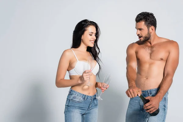 Uomo senza camicia aprendo bottiglia di champagne vicino seducente donna in reggiseno che tiene gli occhiali su grigio — Foto stock