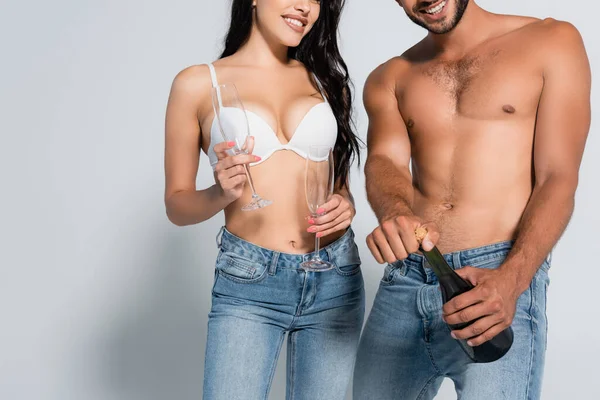 Cropped view of shirtless man opening bottle of champagne near woman in bra holding glasses on grey — Stock Photo