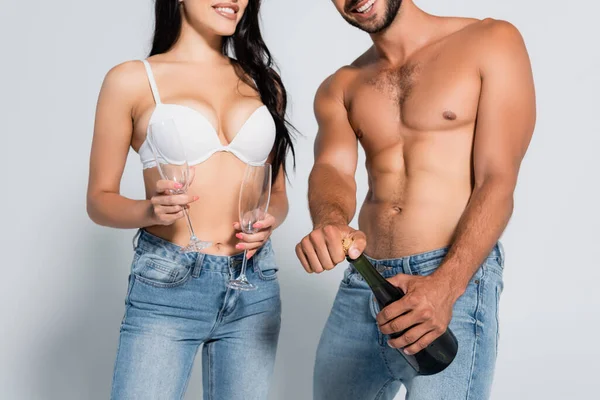 Visão parcial do homem sem camisa abertura garrafa de champanhe perto de mulher sedutora em sutiã segurando óculos em cinza — Fotografia de Stock