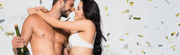 Horizontal image of brunette woman in bra hugging shirtless man with bottle of champagne on grey with confetti — Stock Photo