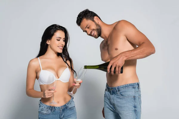 Uomo senza camicia versando champagne in vetro vicino donna sexy in reggiseno su grigio — Foto stock