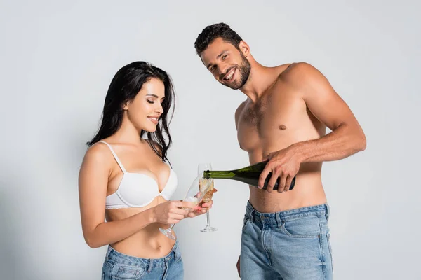 Muskulöser Mann gießt Champagner in Glas neben brünetter Frau in BH auf grau — Stockfoto