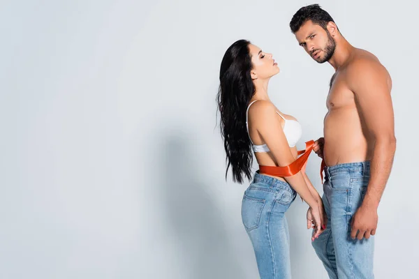 Homme musclé et barbu attacher femme sexy en soutien-gorge et jeans sur blanc — Photo de stock