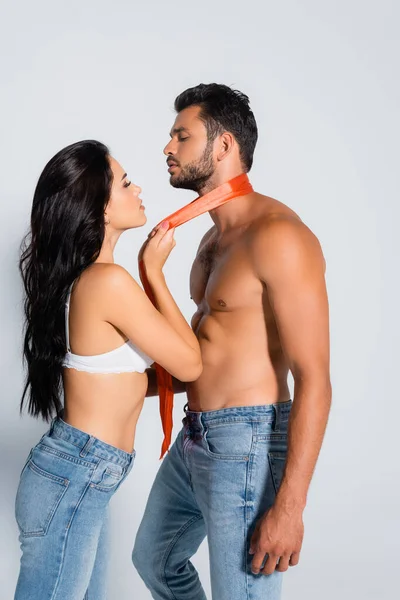 Profile of seductive woman in bra holding ribbon around neck of muscular and submissive man on white — Stock Photo