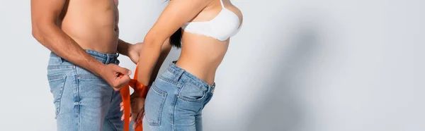 Panoramic crop of muscular man tying sexy woman in bra and jeans on white — Stock Photo