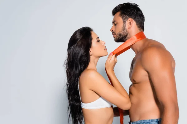 Side view of seductive woman in bra holding ribbon around neck of muscular and submissive man on white — Stock Photo