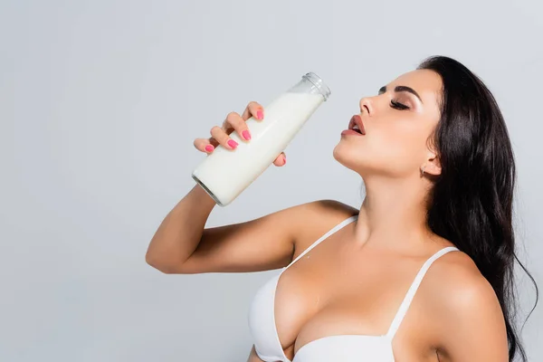 Morena mujer en sujetador sosteniendo botella con leche aislada en gris - foto de stock