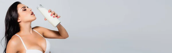 Panoramic crop of sexy woman in bra holding bottle with milk isolated on grey — Stock Photo