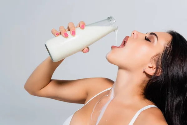 Sexy mujer en sujetador celebración botella y verter leche en boca abierta aislado en gris - foto de stock