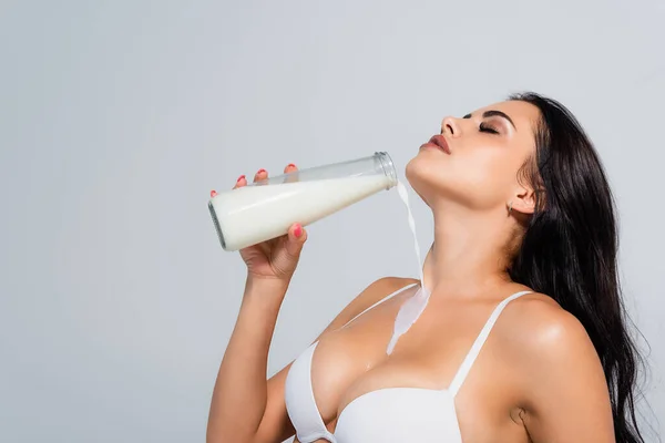 Сексуальная женщина в бюстгальтере с бутылкой и льет молоко на тело, изолированное от серого — стоковое фото