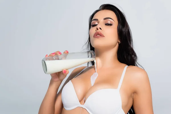 Morena mujer en sujetador celebración botella y verter leche en busto aislado en gris - foto de stock