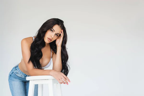 Sexy junge Frau in BH und Jeans lehnt auf Stuhl und blickt in die Kamera auf grau — Stockfoto