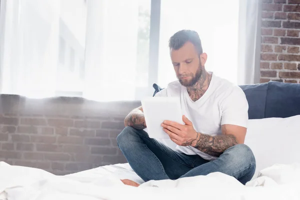 Tattooed man in white t-shirt and jeans using digital tablet while sitting in bed with crossed legs — Stock Photo