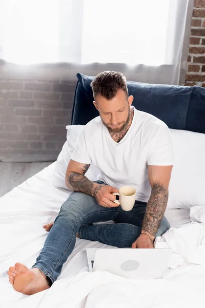 Tattooed freelancer in white t-shirt and jeans using laptop in bed while holding cup of tea — Stock Photo