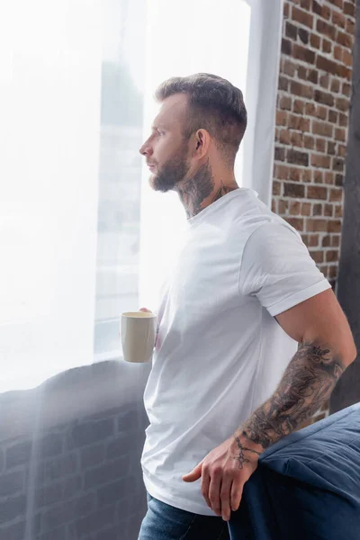 Вид сбоку на молодого человека с татуировкой, который смотрит в сторону, стоя у окна с чашкой чая — стоковое фото