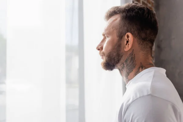 Side view of bearded, tattooed man looking away while standing near window at home — Stock Photo