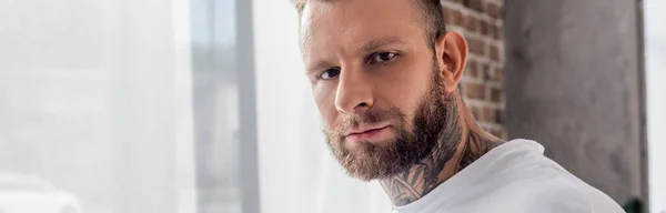 Panoramic crop of confident, tattooed man looking at camera near window — Stock Photo