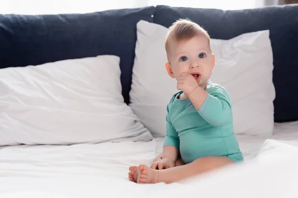 Selektiver Fokus des Säuglings im Babystrampler, der mit der Hand im Mund im Bett sitzt — Stockfoto