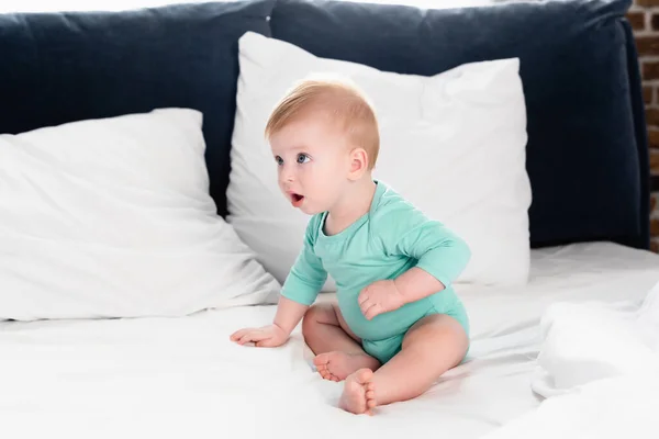 Menino em bebê romper sentado na cama e olhando para longe com a boca aberta — Fotografia de Stock