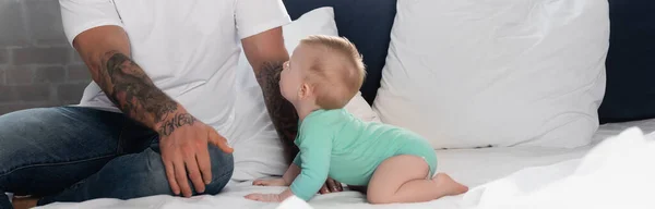 Panoramic concept of infant boy in baby romper crawling on bed near father — Stock Photo