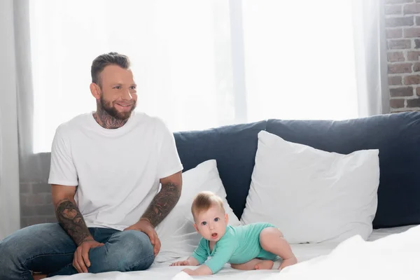 Excited young man in white t-shirt and jeans sitting near baby boy crawling on bed with open mouth — Stock Photo