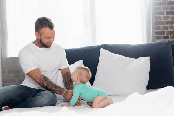 Young man in white t-shirt and jeans touching infant son crawling on bed in baby romper — Stock Photo