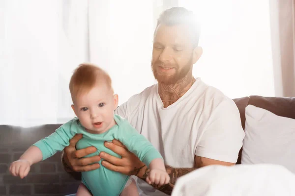Enfoque selectivo de joven hombre tatuado sosteniendo bebé niño mientras está sentado en el dormitorio - foto de stock