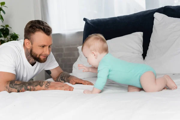 Selektiver Fokus tätowierter junger Mann hat Spaß mit Säugling, der im Babystrampler auf Bett krabbelt — Stockfoto