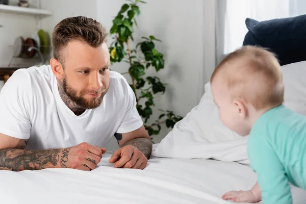 Foco seletivo de homem tatuado olhando para criança infantil rastejando na cama — Fotografia de Stock