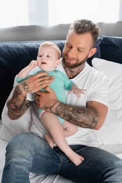 Giovane uomo barbuto in jeans tenendo bambino ragazzo mentre seduto a letto — Foto stock