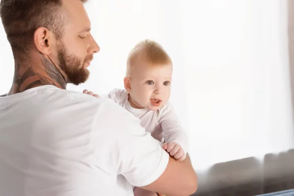 Niño mirando hacia otro lado con la boca abierta en las manos en el joven padre tatuado - foto de stock