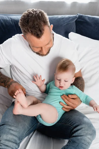 Padre joven en camiseta blanca y jeans sentados en la cama y divirtiéndose con su hijo pequeño - foto de stock