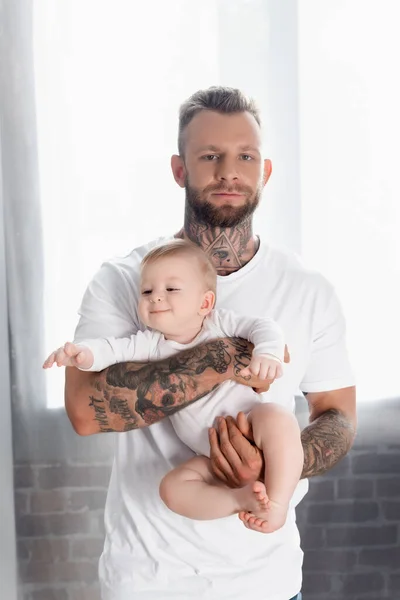 Junger tätowierter Mann blickt in die Kamera, während er seinen fröhlichen kleinen Sohn im Babystrampler hält — Stockfoto