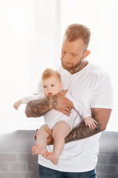 Jovem barbudo homem no branco t-shirt segurando criança infantil no bebê romper — Fotografia de Stock