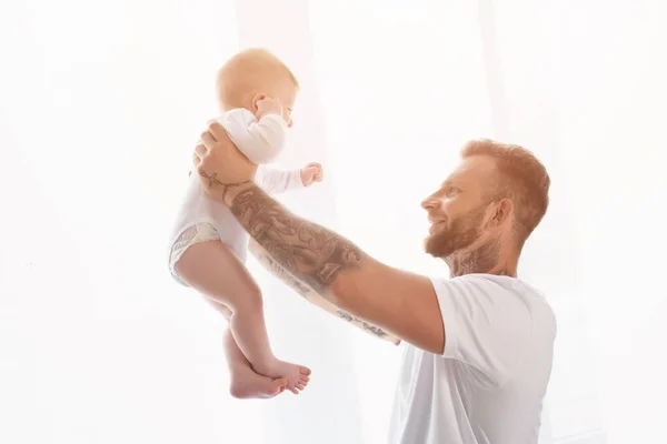 Gioioso uomo tatuato in t-shirt bianca che tiene il figlio neonato in mani tese — Foto stock